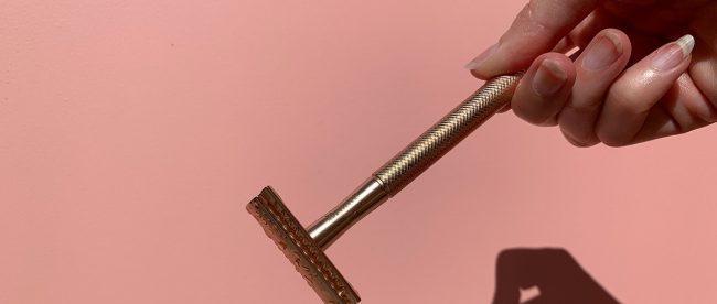 Hand holding a razor on a plain pink background, preparing to throw it away