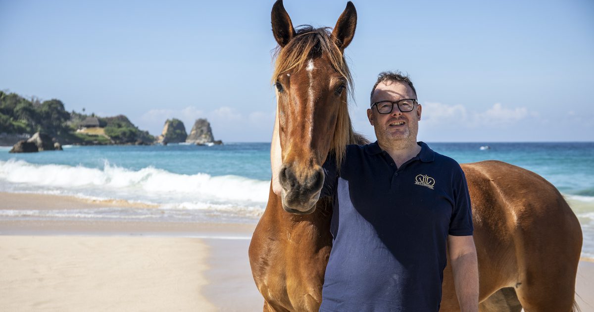 Man stood in front of a brown horse on the beach with a blue sea behind them. 