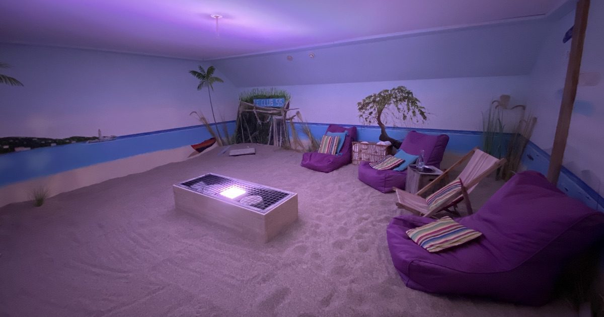 A photo showing the Sabbia Med room. The floor is covered in sand, the walls painted to look as though you're sitting at the beach. The room has deck chairs and slouching cushions to sit on, and low level lighting