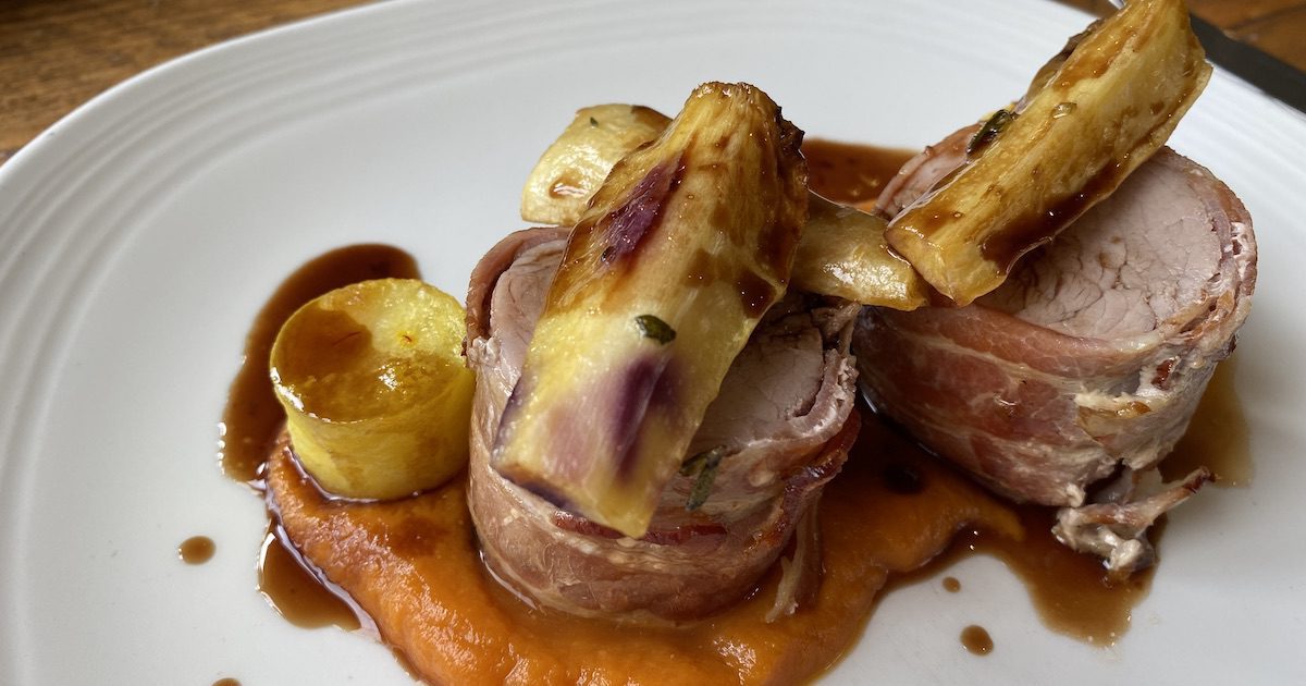 Plate shows small bundles of pork fillet wrapped up in pancetta on a bed of carrot puree and with roasted root vegetables on top