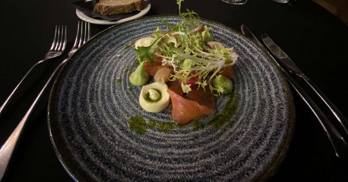 Image shows plate with smoked salmon, small dollops of different sauces and oils, some frisse lettuce leaves, with brown bread on a plate behind.