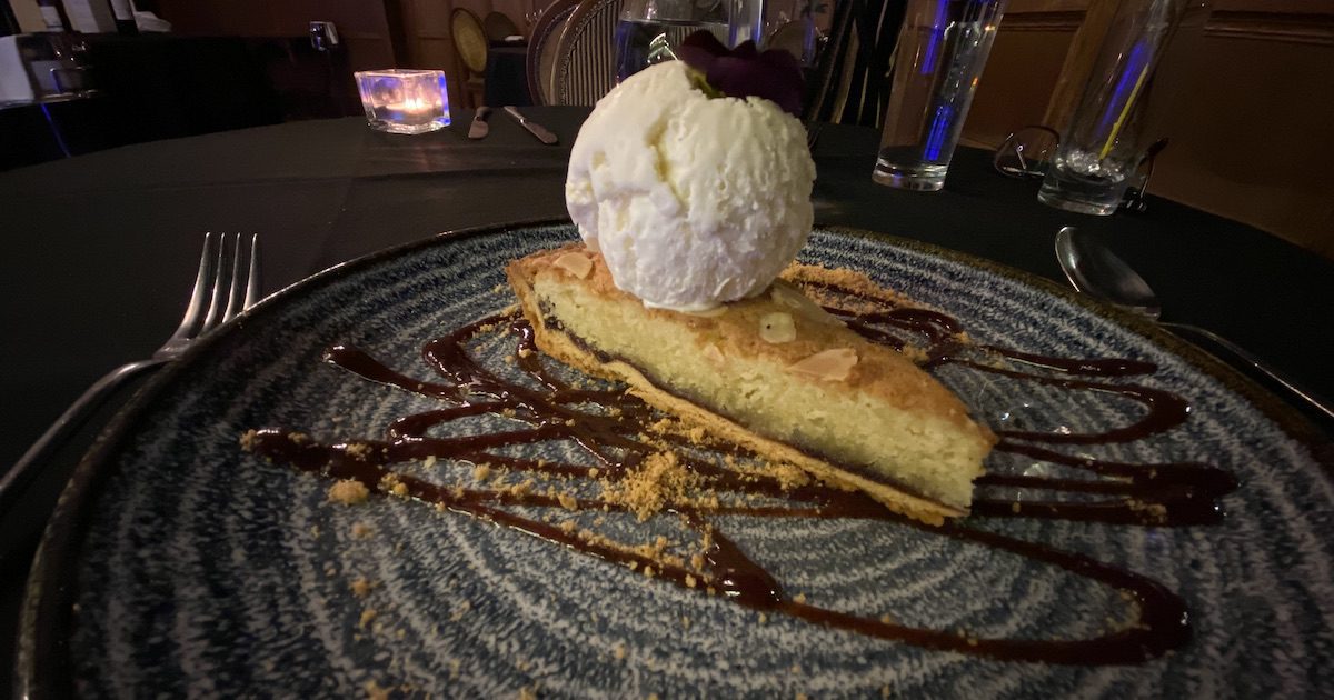 Plate showing slice of warm blackberry bakewell tart and large dollop of vanilla ice cream, with sauce drizzled around