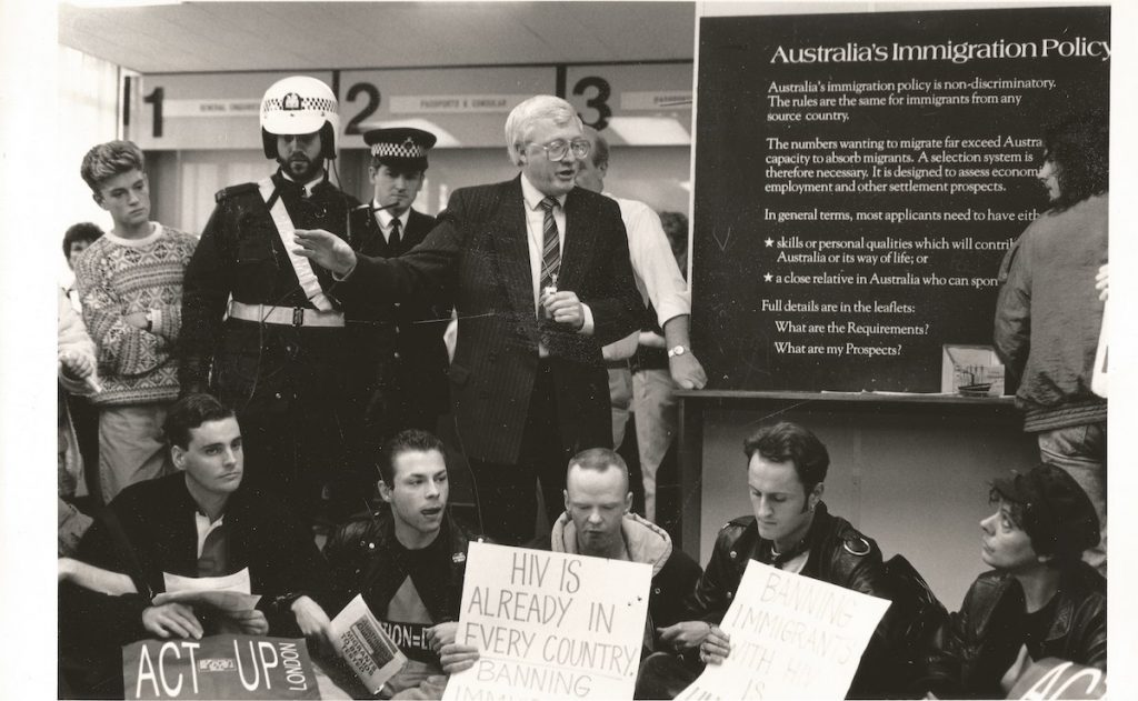 Black and white photo of members of ACT UP in Australia knelt in front of a board titled 'Australia's Immigration Policy' holding a sign that says 'ACT UP London'