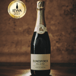 Dunesforde Blanc de Noir in a dark bottle with gold foil top and white label across the middle, set against a brown background with a gold sticker in the top left corner for the wine's gold award in 2021 in the IEWA