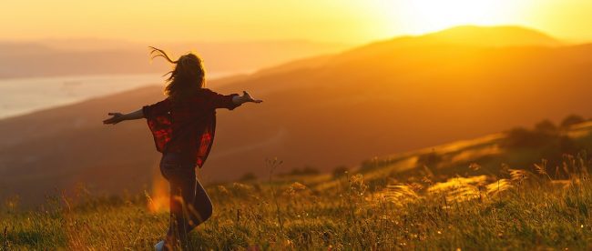 Image shows woman happy and carefree feeling awestruck by a beautiful sunset, arms raised