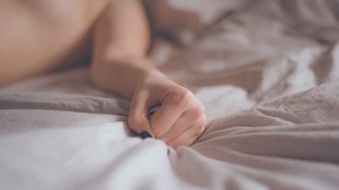 Image shows a hand grabbing a sheet on the bed, body is out of focus, demonstrating sexual pleasure