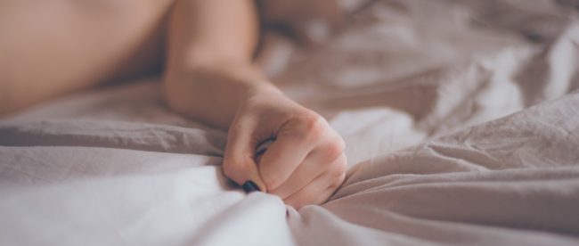 Image shows a hand grabbing a sheet on the bed, body is out of focus, demonstrating sexual pleasure