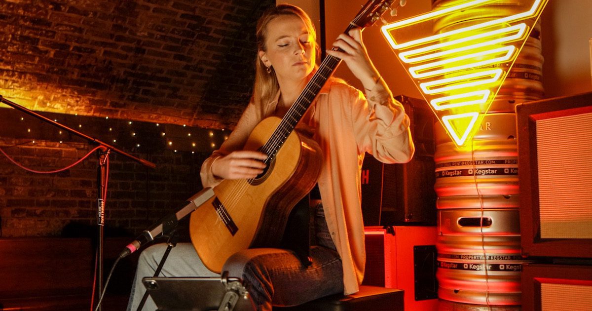 A blond woman sat playing acoustic guitar behind a neon yellow triangle sign. Part of a classical club night noisenights from Through the Noise