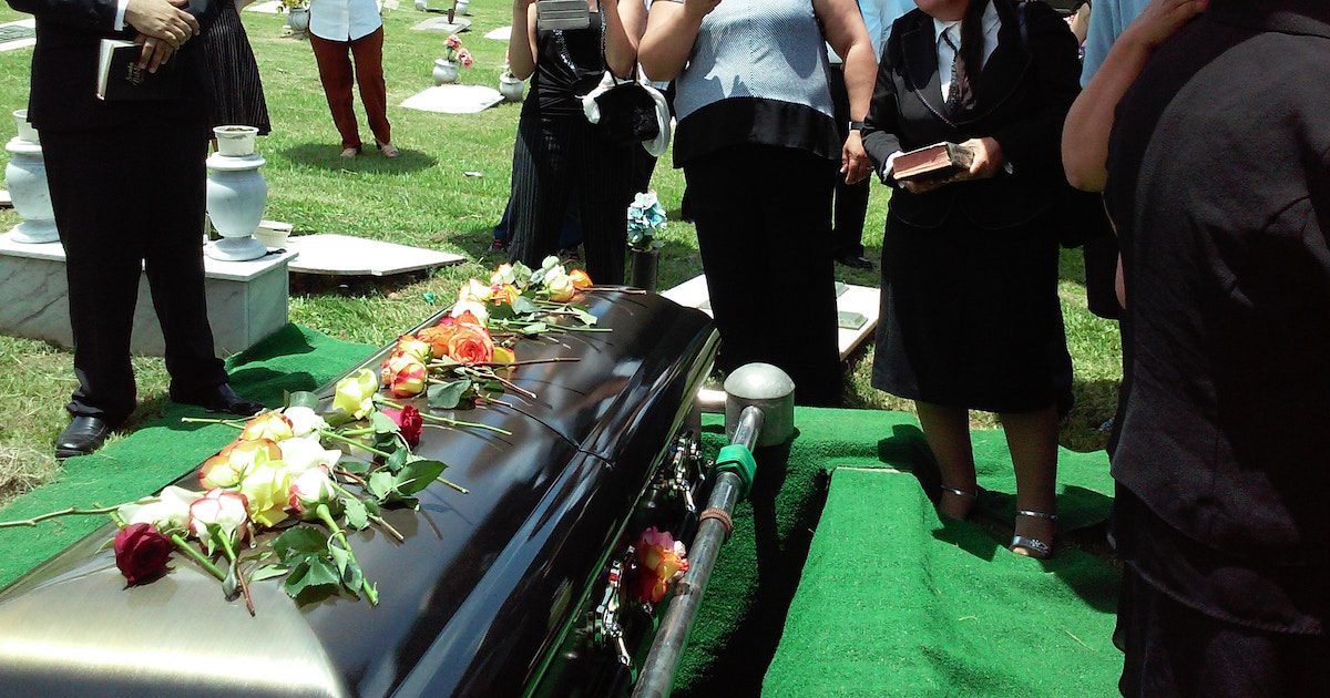 People stood around a black coffin, outside, with flowers scattered across it. The psychology behind funeral crashers