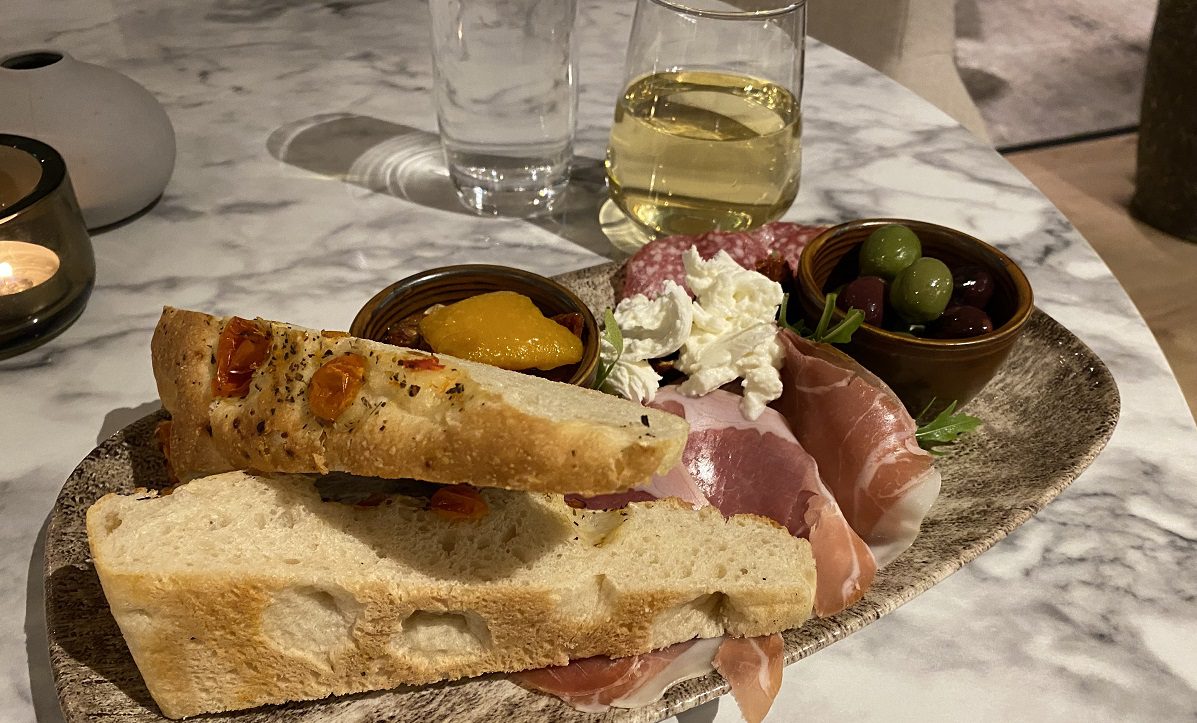 Image shows a plate of meats, cheese, olives, bread etc, and a glass of wine in the background