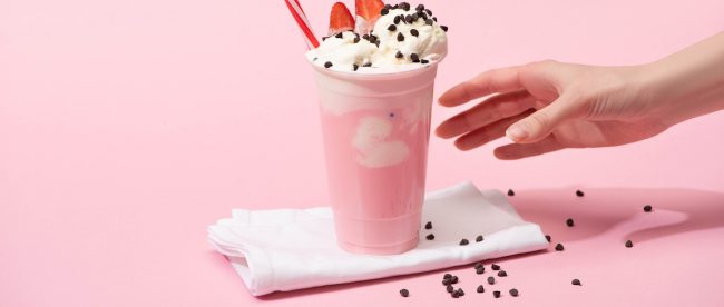 Partial view of female hand with disposable cup of milkshake with chocolate morsels and strawberry on napkins on pink.