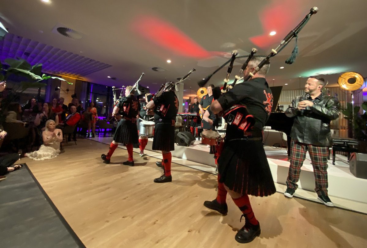 Image shows a band of pipers in black and red kilts with bagpipes