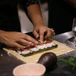 Someone rolling out sushi, filled with vegetables, on a wooden board.