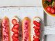 Four eclairs decorated with pink icing and strawberries on an off-white wooden chopping board next to a bowl of strawberries. Strawberry and raspberry eclair recipe.