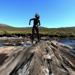 Woman dancing on rock in wetsuit by the sea.