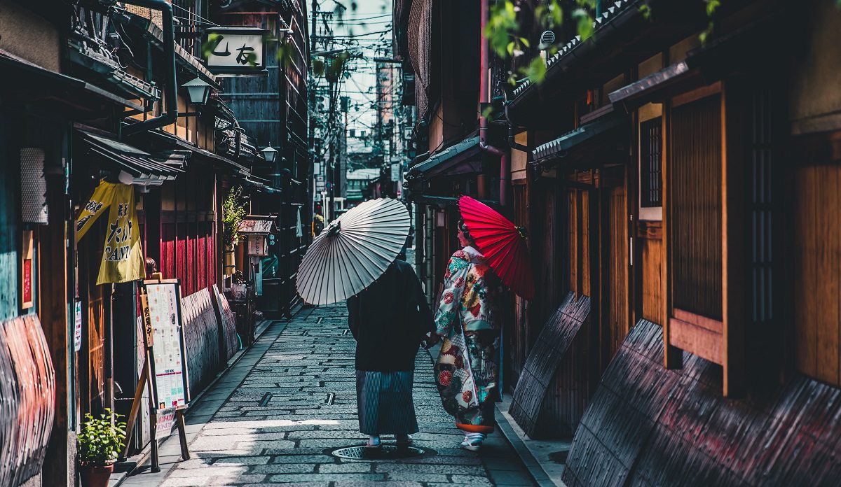 Image shows two Japanese women walking away from the shot dressed in traditional clothing, in a traditional Japanese street in Kyoto