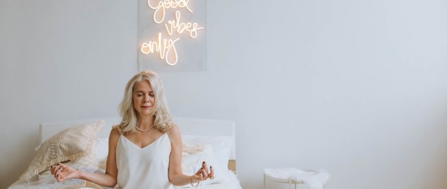 Woman sat in white pyjamas meditation on bed with 'Good Vibes Only' light up sign behind her