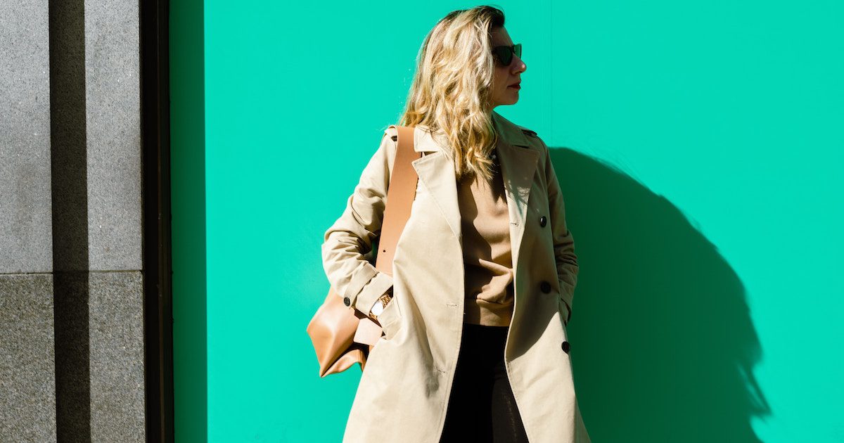 Woman stood in front of green wall wearing beige trench coat. Autumn wardrobe staples