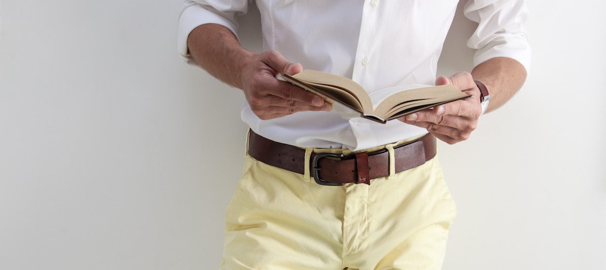 Man's waist reading a book. Wearing pale yellow trousers, leather belt, and a white shirt. Wardrobe essentials autumn.