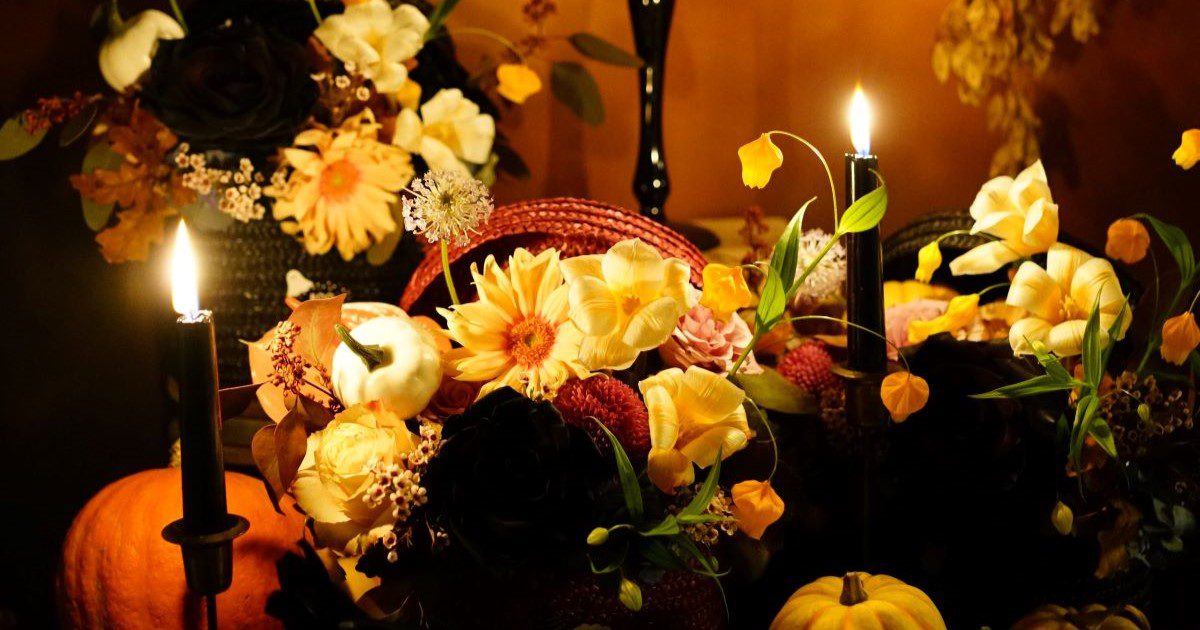 An arrangement of yellow flowers and miniature pumpkins spilling from a basket with three lit black candles surrounding it