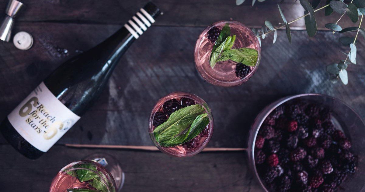 A bowl of blackberries and a laid down bottle of alcohol sit either side of three glasses, each filled with a rich purple blackberry drink