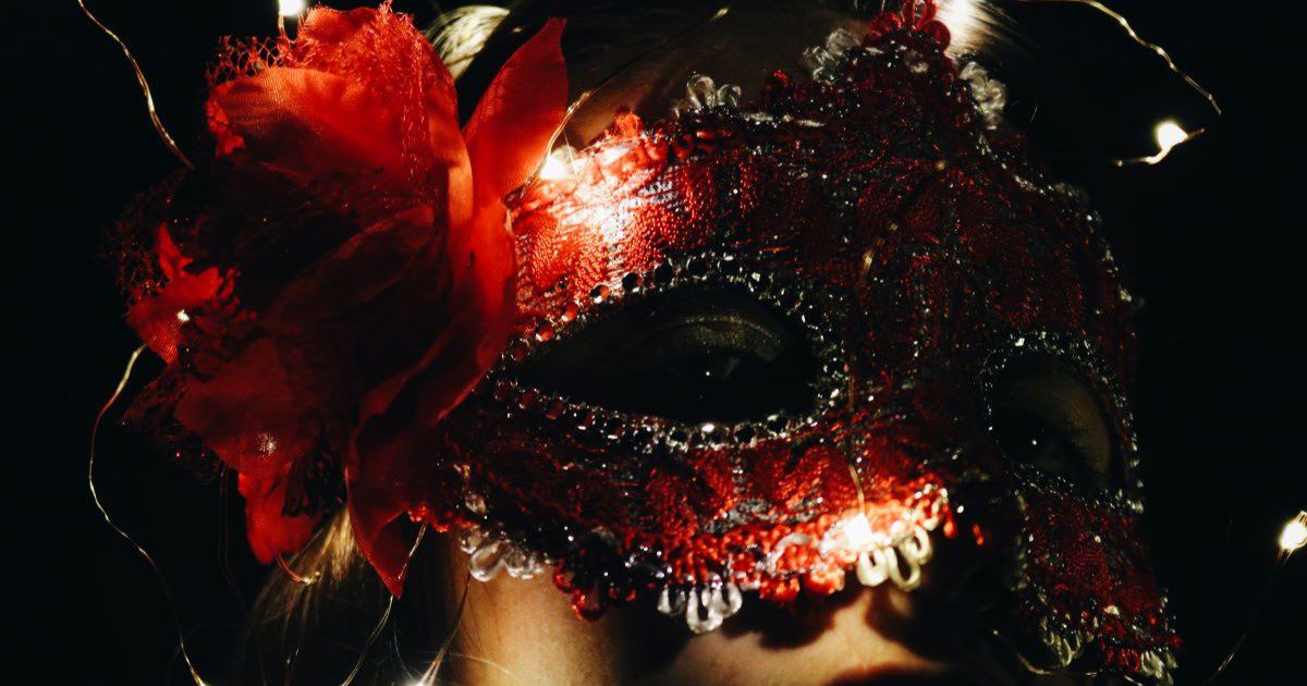 A dark image of a person wearing an extravagent red masquerade mask with a red flower attached