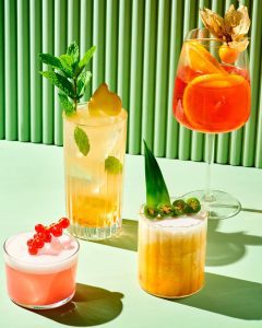Examples of cocktails available at Torstigbar. Image shows an array of yellow, orange and pink fruity cocktails decorated simply with fruits and leaves, all on a simple green backdrop.