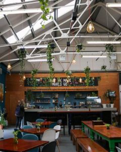 Inside of The Brink. There are wooden tables with benches and white and blue chairs surrounding them; a simple bar at the back with a chalkboard-looking sign that is obscured by leaves. It reads "The Brink. What we make makes us". Steel beams and green plants cover the ceiling.