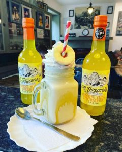 Inside of Mr Fitzpatrick's Temperance Bar, showing a drink from their menu. A mason jar glass filled with a thick lemon milkshake, topped with whipped cream and a Party Ring with a striped red and white straw through it. There are two bottles of lemon, yuzu and tumeric cordial beside it, both branded as Mr Fitzpatrick's.