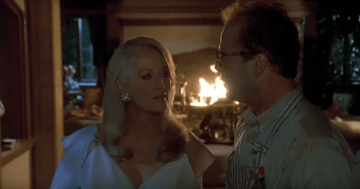 A young, blonde, Meryl Streep stood in front of a fire facing a dark haired man wearing glasses. Death Becomes Her a Halloween film not scary idea