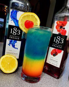 An example of YADA Collective's no alcohol cocktails. A multicoloured cocktail, blue on top, then a yellow to red gradient beneath, topped with a lemon slice and a cherry. Beside the glass are two bottles of alcohol and a second lemon slice.