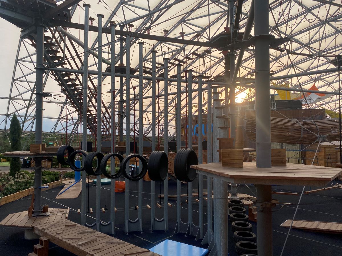 An image showing the inside of Bluestone Resort's Serendome. It has huge structures to climb on, including a series of hung up tires, high up stairs, wires, and more. The ceiling is mostly glass, with a view of the sky and a low set sun.