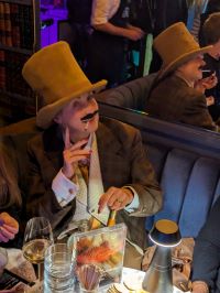 Image of a person with a tall yellow tophat, wearing a brown suit and yellow tie. They have a fake moustache and red nail polish. They are looking at the camera, pointing a finger upwards with squinted eyes, a smile, and an unlit cigarette in their mouth.