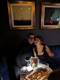 Image of a man dressed in all black, with thick black rimmed glasses. A woman is sat beside him, dressed in a black dress and a pearl necklace, with tied back black hair. The two are leant against each other, smiling, with a cheese platter on the table in front of them.