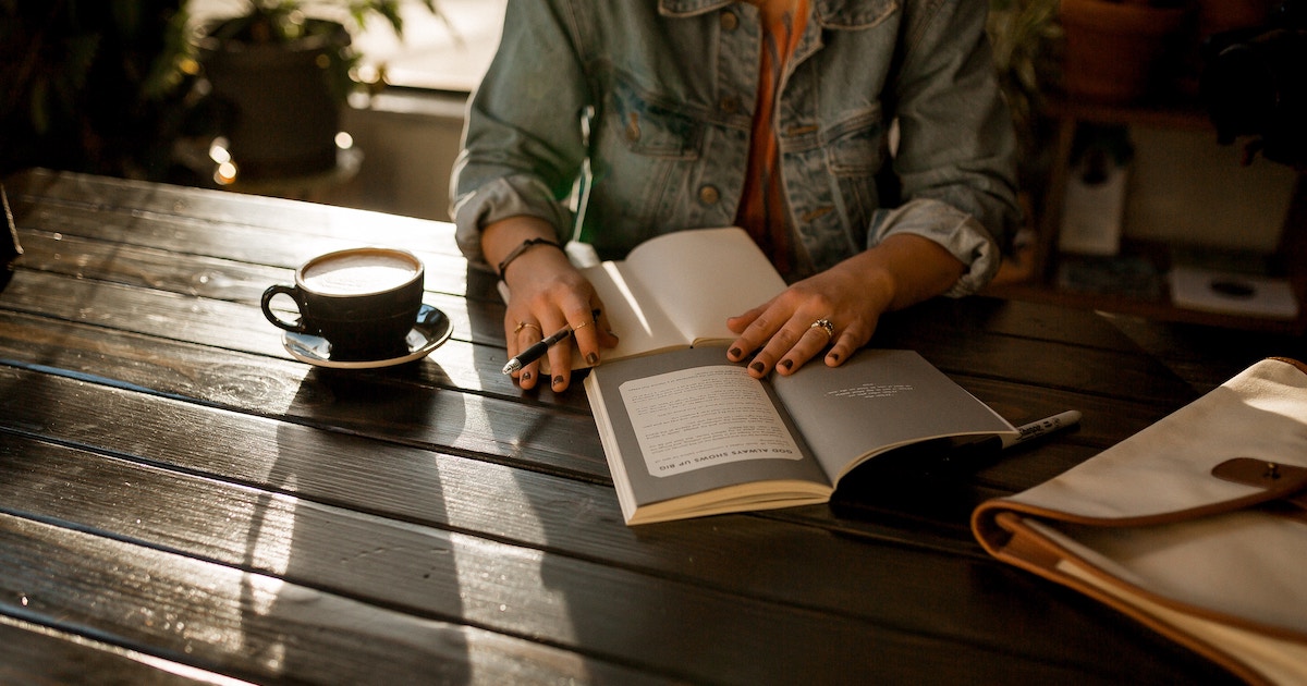 Person sat, wearing a denim jacket, journalling in a cafe with a cup of coffee. Learn the benefits of journalling on Silver