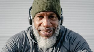 Mature man with grey beard wearing a dark green beanie hat and headphones smiling into the camera. Men's health looking and feeling better