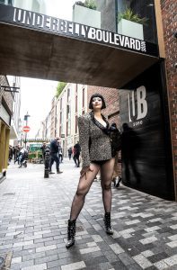 An image of Bernie Dieter stood outside of the Underbelly Boulevard. She has black hair in a short and straight bob cut, and is wearing a short dress which resembles a business suit, with large shoulder pads, as well as fishnets. She is posed with her leg out, hand on her thigh, looking towards the camera.