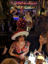 Darkened image of a woman with a wide brimmed mushroom hat and a floral red dress. She is chatting with a group of people in a bar, with a mirror behind reflecting the scene.