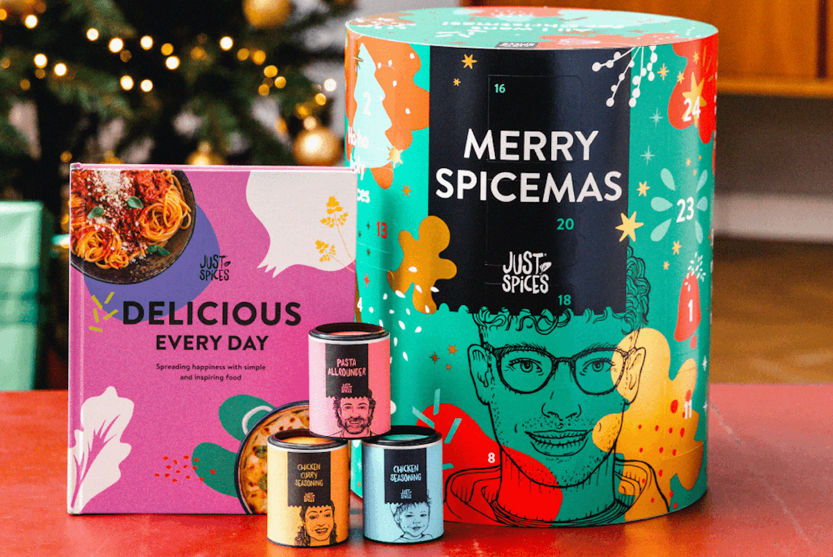 Cylindrical tub in teal with 'Merry Spicemas' written on the front and small tubs of spices sat next to it. All on a table in front of a decorated Christmas tree.