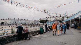 Lots of people walking and standing along a pier, with bunting overhead and terraced housing in the background.
