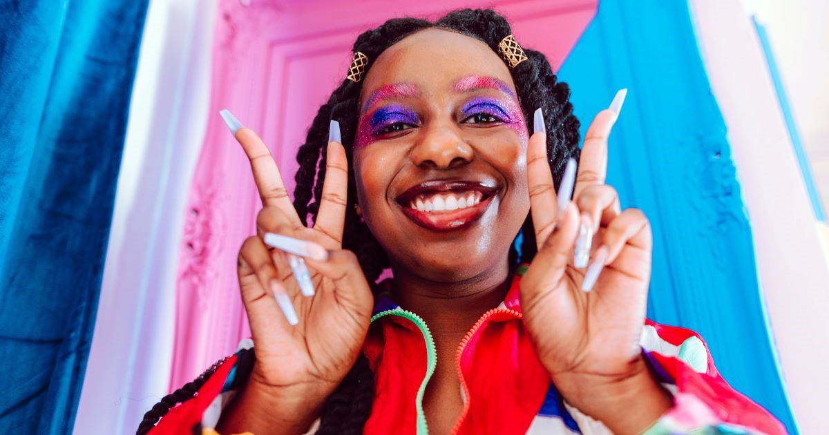 Image of a woman with a wide smile, holding up two peace signs. She has long black hair in dreads, a red jacket and blue, pink and purple makeup. The background is the colours of the transgender pride flag - blue, pink and white.