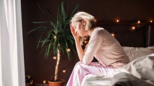 Image of a woman sat on the edge of her bed. She is holding her head in one hand, looking stressed.