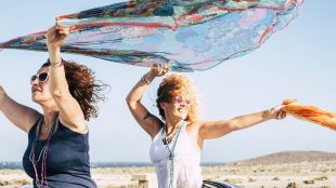 Couple of women have fun and play with the wind standing out of the top of a convertible car and playing with coloured pareos - cheerful people traveling and enjoying the outdoor on vacation