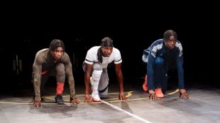 Three young black men dressed in sports gear are lined up as though ready to run a race, in a scene from the play